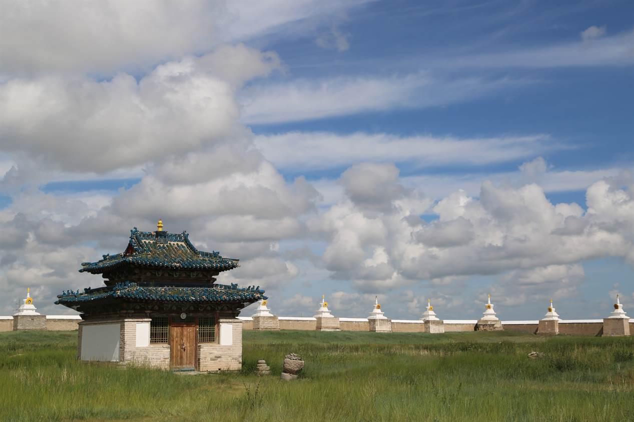 A traditional Mongolian building sits in a field in front of a brick wall with decorative spires