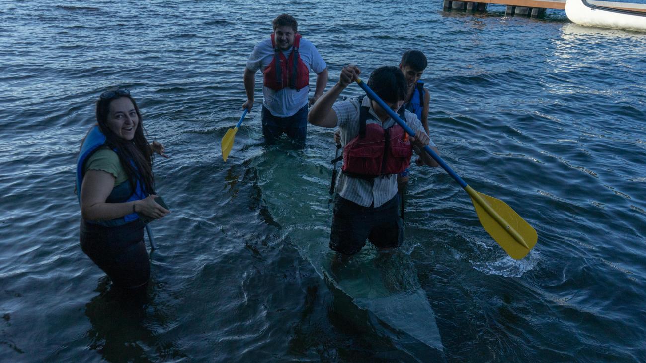 people standing in the water with life jackets and boat in water, person in front is holding an oar