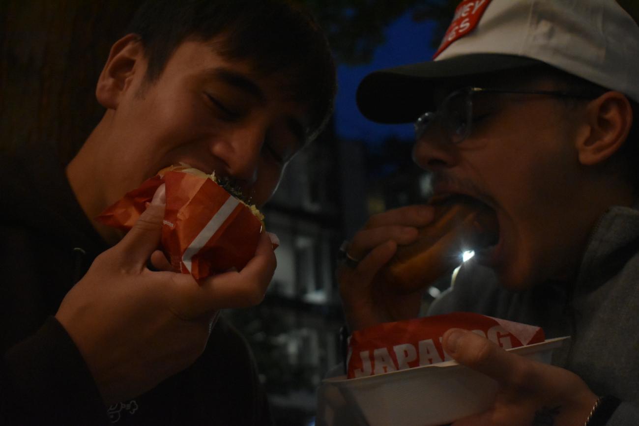two students eating hot dogs, while it is dark and they are outside