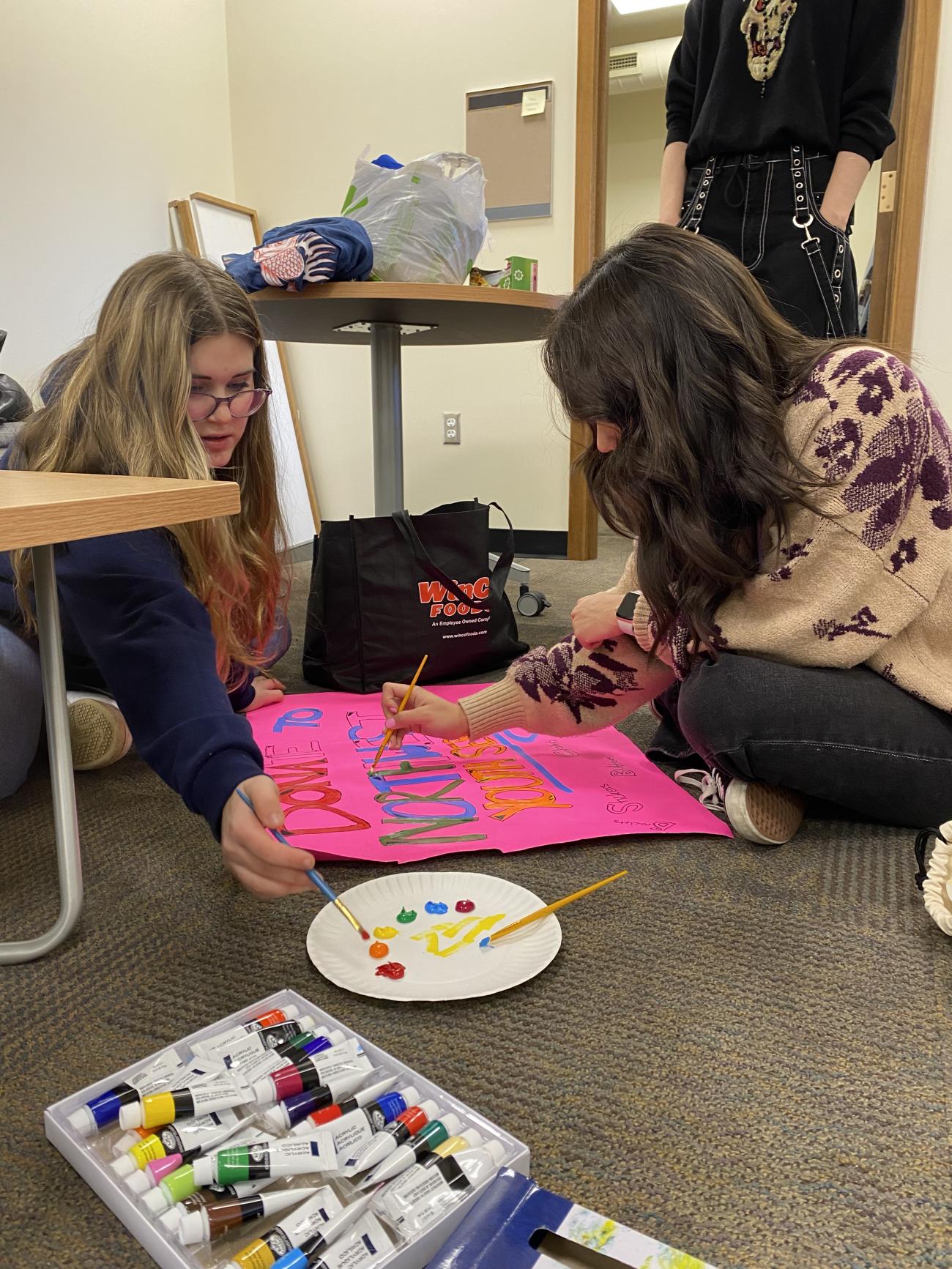 two students sitting on the floor using paint to decorate a poster