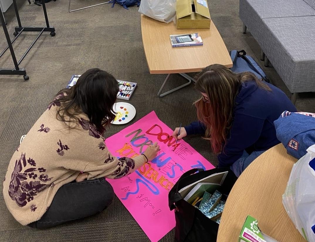 two students sitting on the floor of a classroom decorating a poster