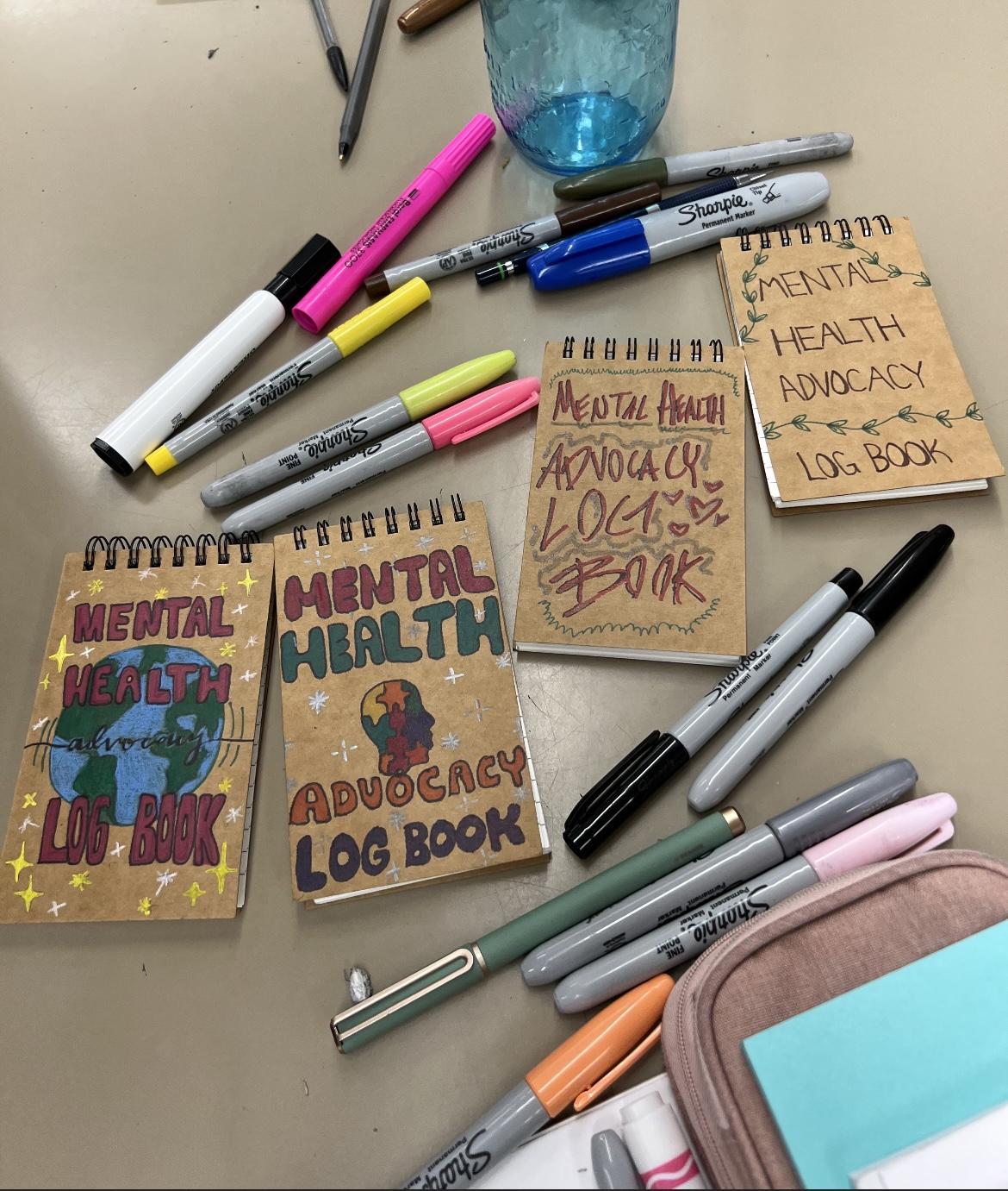 notebooks decorated with mental health designs on the cover with pens around the notebooks