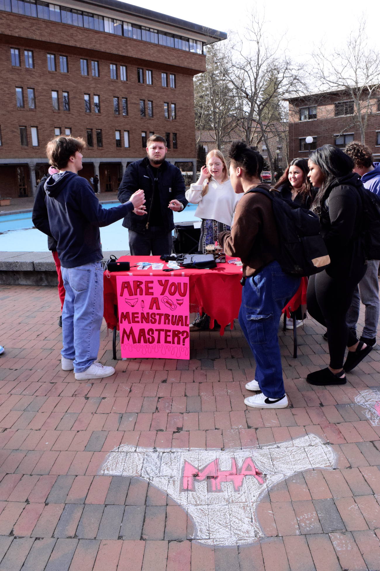 People cstanding near table decorated with red tableclothe and posters, and chalk drawing on brick ground