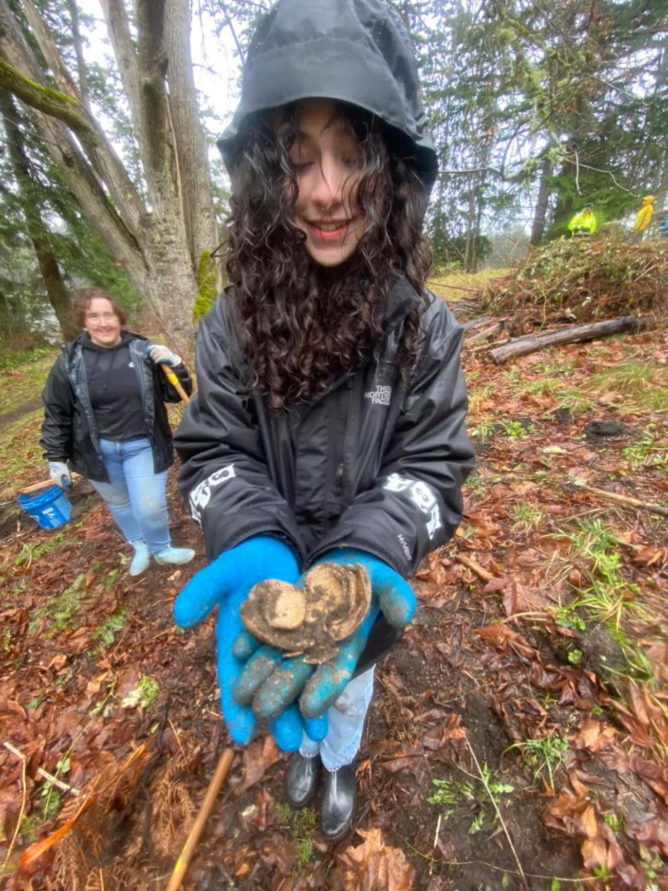person in the woods, wearing gloves, holding something and looking down at it, showing the camera