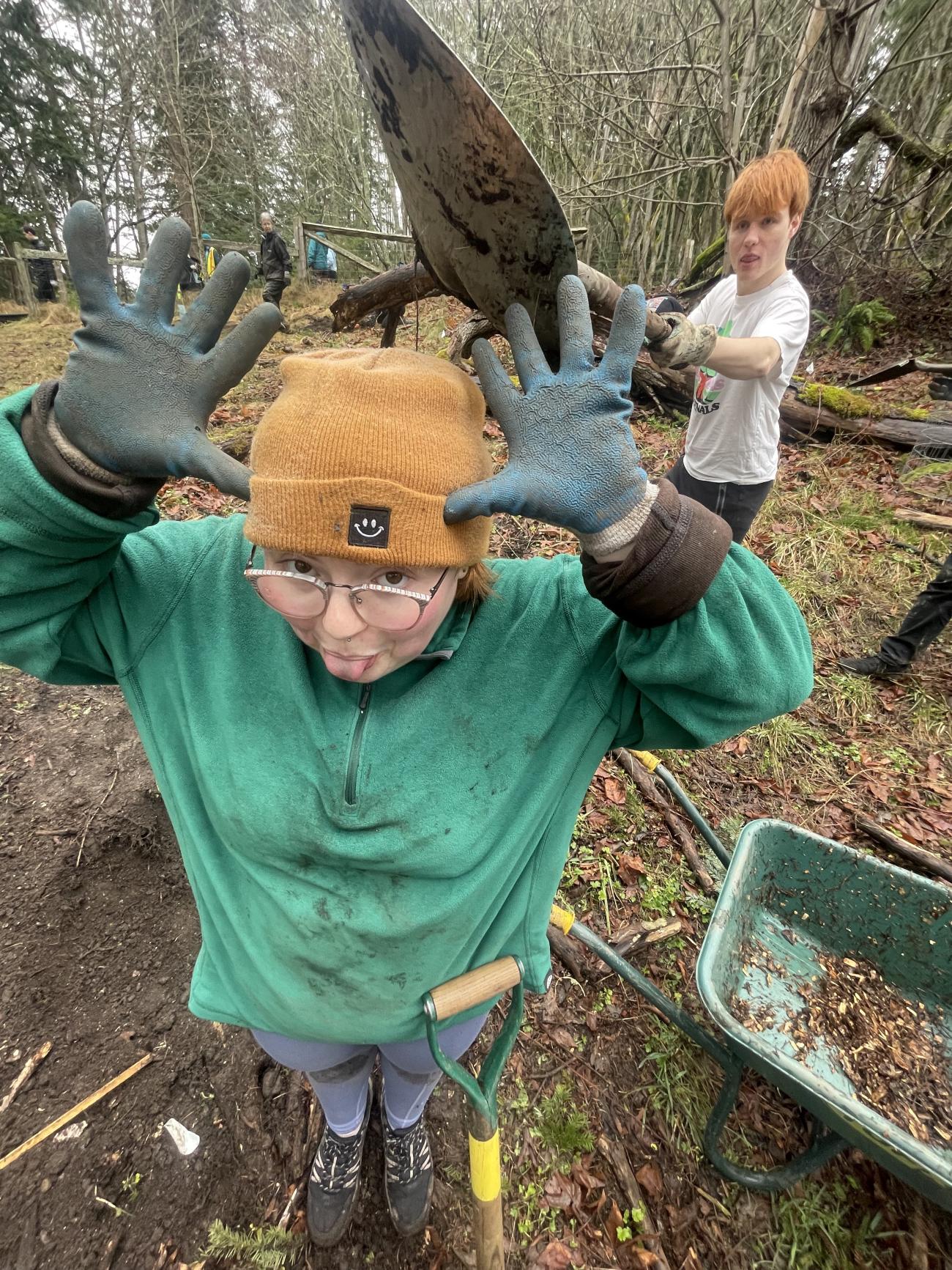 person with hands spread, thumbs next to ears, sticking their tongue out, while someone behind them holds a shovel over the person's head