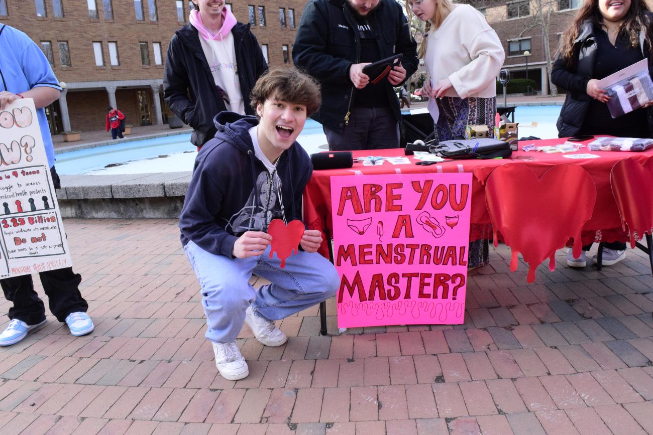 person squatting and smiling infront of table while holding a red paper heart in front of them, next to a pink sign that reads 'are you a menstrual master?'