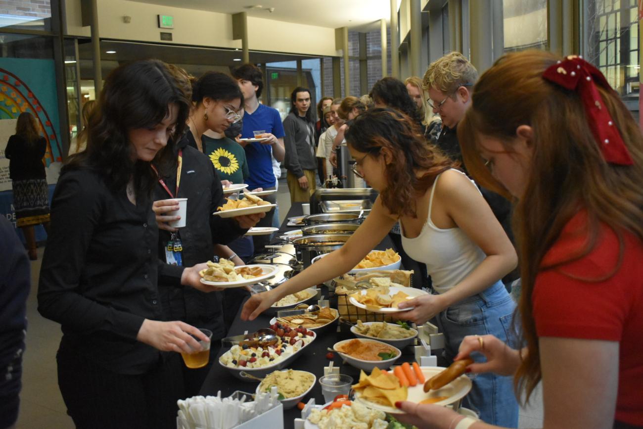 students grabbing food and putting them on paper plates, from a  long table with black table clothe and multiple different dishes